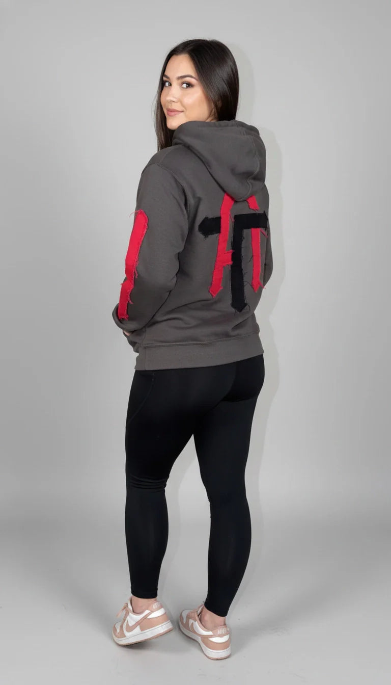 A woman wearing the True Authentic charcoal grey streetwear hoodie with deconstructed red and black patchwork on the back and sleeves, paired with black leggings and sneakers against a clean studio background.
