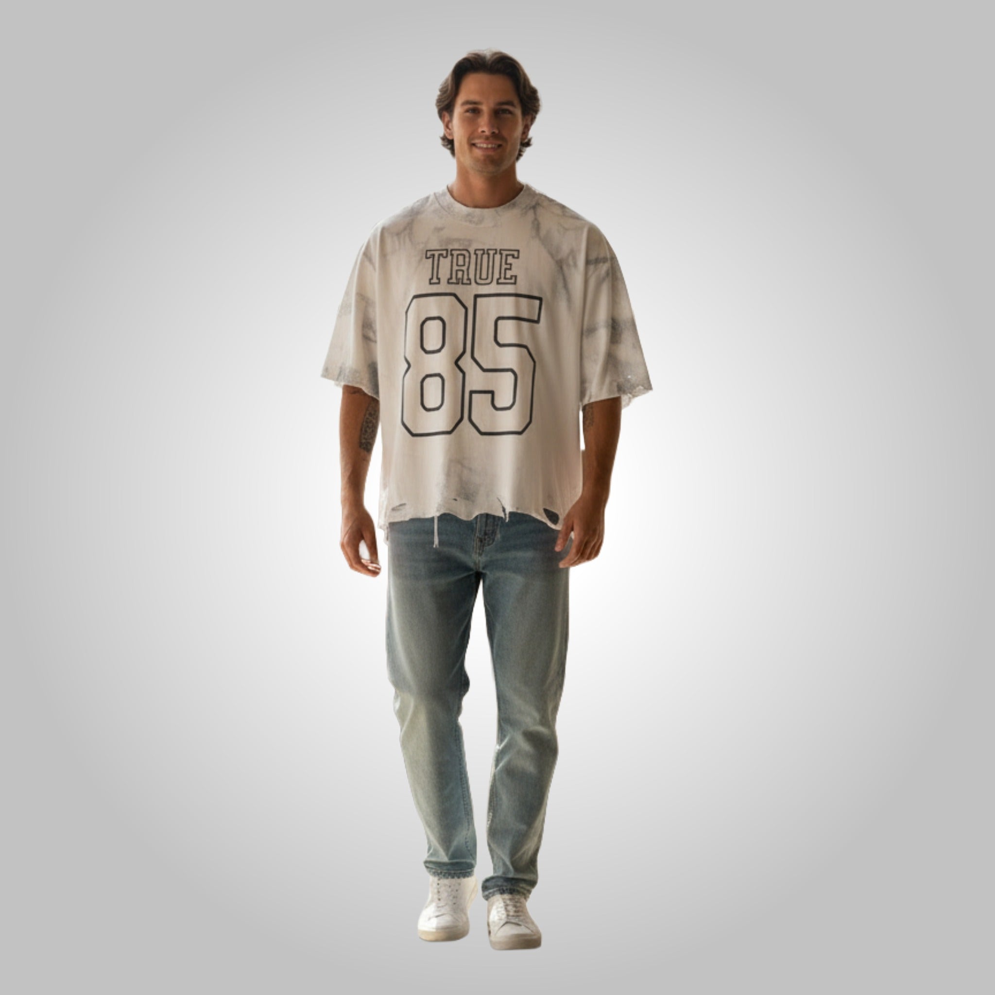 A male model wearing a white deconstructed "True 85" oversized t-shirt with a vintage collegiate design and light-wash jeans.