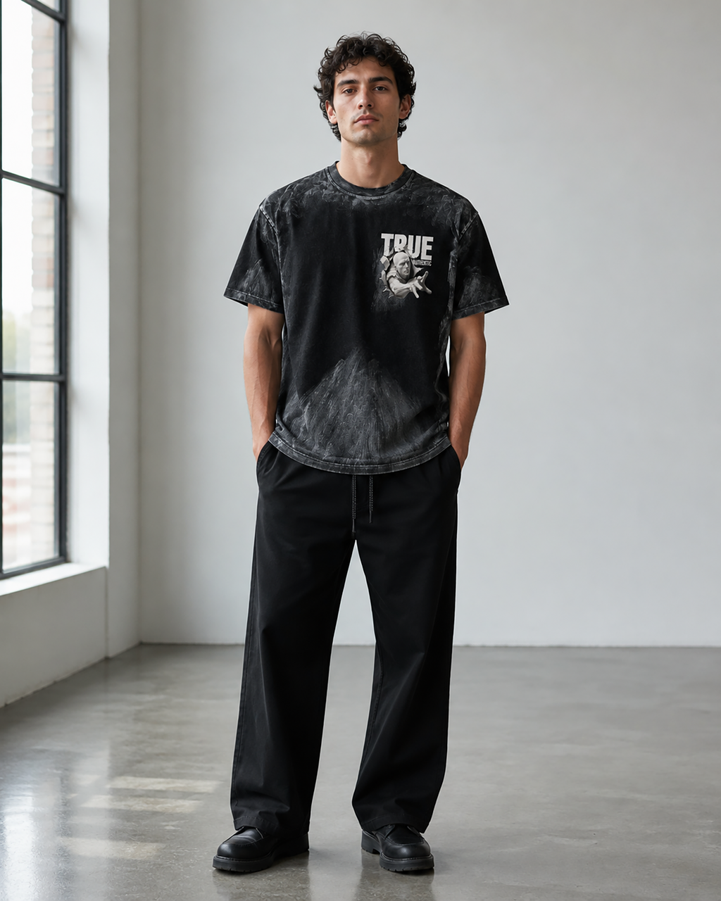 A man stands in a bright studio wearing a black acid-wash graphic t-shirt with 'TRUE' and a skull graphic on the chest, paired with black sweatpants and black shoes.