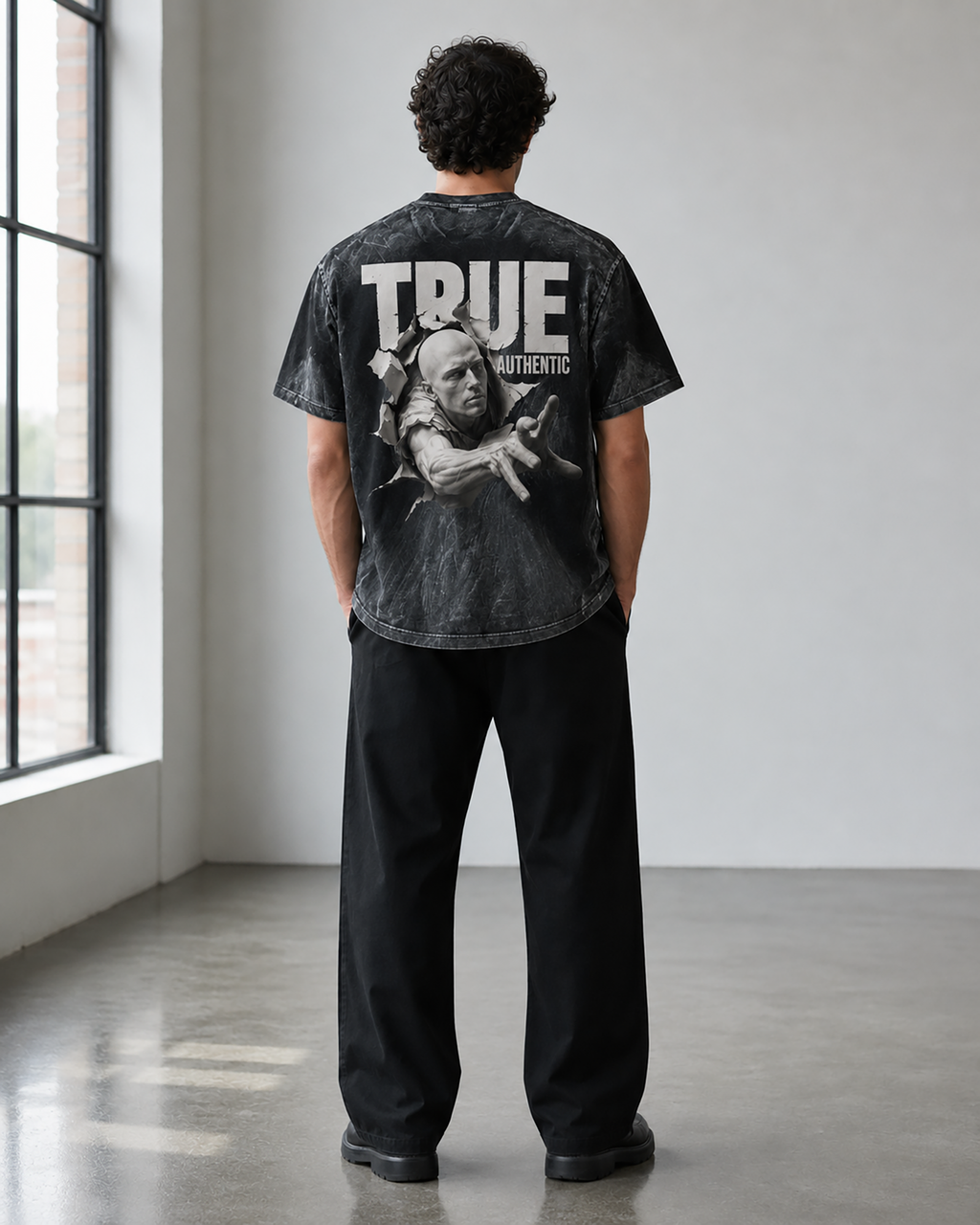 A man stands with his back to the camera wearing a faded black graphic t-shirt with 'TRUE AUTHENTIC' and a figure printed on the back, paired with black pants in a minimalist studio setting.