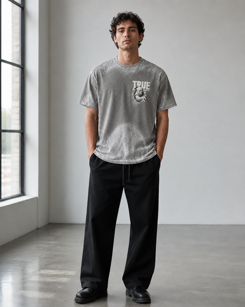 A man wearing a heather gray crewneck t-shirt with a distressed graphic on the chest that reads 'TRUE' and shows a monkey, paired with black wide-leg trousers, standing in a minimalist studio with a large window.