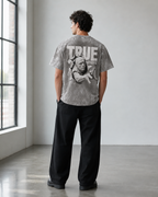 A man stands with his back to the camera in a bright studio, wearing a gray acid-wash graphic t-shirt with a large 'TRUE GUY' print featuring a bald head illustration, paired with black pants.