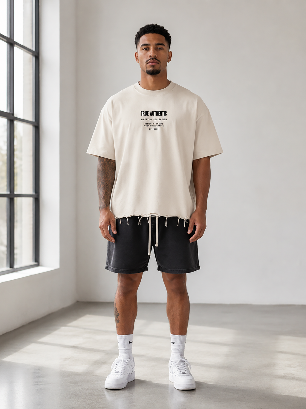 A man modeling a cream-colored t-shirt with black graphic text on the chest, paired with black drawstring shorts, white socks, and white sneakers in a bright, modern room with a large window.