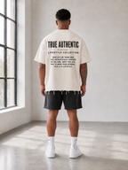 A man stands with his back to the camera in a bright, minimalist studio, wearing a cream-colored t-shirt with black text on the back reading 'TRUE AUTHENTIC LIFESTYLE COLLECTION', black shorts, white socks, and white sneakers.