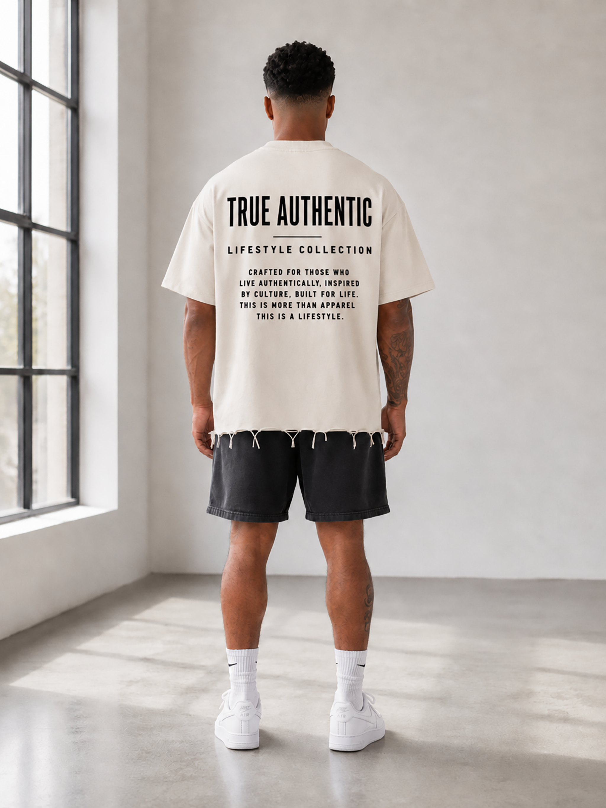 A man stands with his back to the camera in a bright, minimalist studio, wearing a cream-colored t-shirt with black text on the back reading 'TRUE AUTHENTIC LIFESTYLE COLLECTION', black shorts, white socks, and white sneakers.
