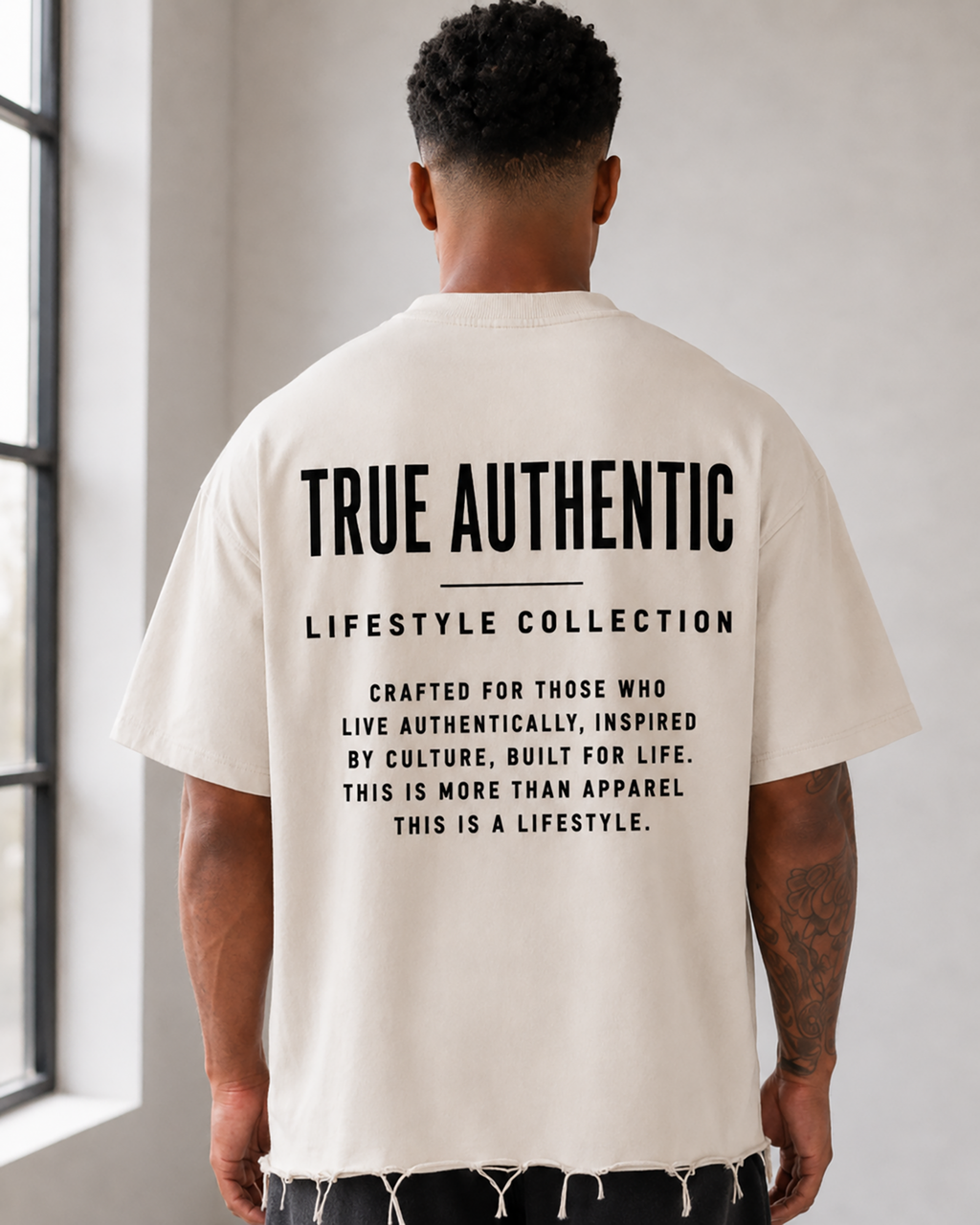 A man stands with his back to the camera in a bright, minimalist studio, wearing a cream-colored t-shirt with black text on the back reading 'TRUE AUTHENTIC LIFESTYLE COLLECTION', black shorts, white socks, and white sneakers. close up