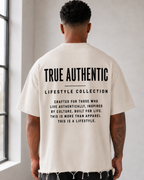 A man stands with his back to the camera in a bright, minimalist studio, wearing a cream-colored t-shirt with black text on the back reading 'TRUE AUTHENTIC LIFESTYLE COLLECTION', black shorts, white socks, and white sneakers. close up