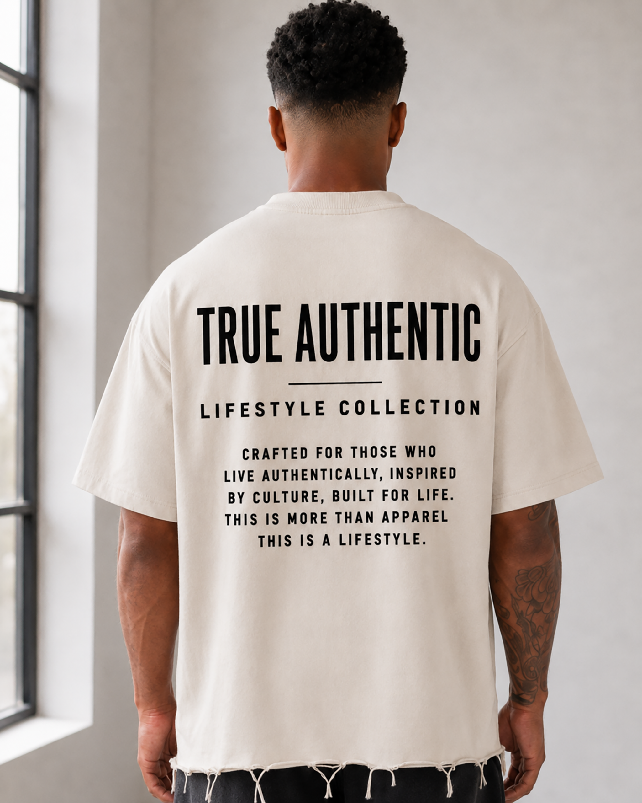 A man stands with his back to the camera in a bright, minimalist studio, wearing a cream-colored t-shirt with black text on the back reading 'TRUE AUTHENTIC LIFESTYLE COLLECTION', black shorts, white socks, and white sneakers. close up