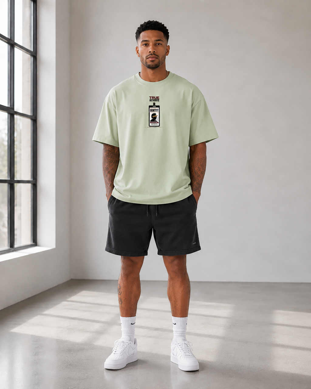 A man stands in a bright studio wearing a light sage green t-shirt with a black graphic print on the chest, black athletic shorts, white socks, and white sneakers.