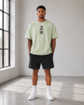 A man stands in a bright studio wearing a light sage green t-shirt with a black graphic print on the chest, black athletic shorts, white socks, and white sneakers.
