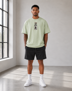 A man stands in a bright studio wearing a light sage green t-shirt with a black graphic print on the chest, black athletic shorts, white socks, and white sneakers.