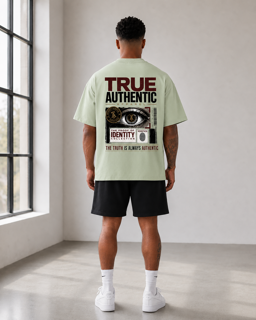 A light sage green short-sleeve t-shirt shown from the back, featuring a large graphic print with the text 'TRUE AUTHENTIC APPAREL' and a collage of an eye, a fingerprint, and security badge elements. male model