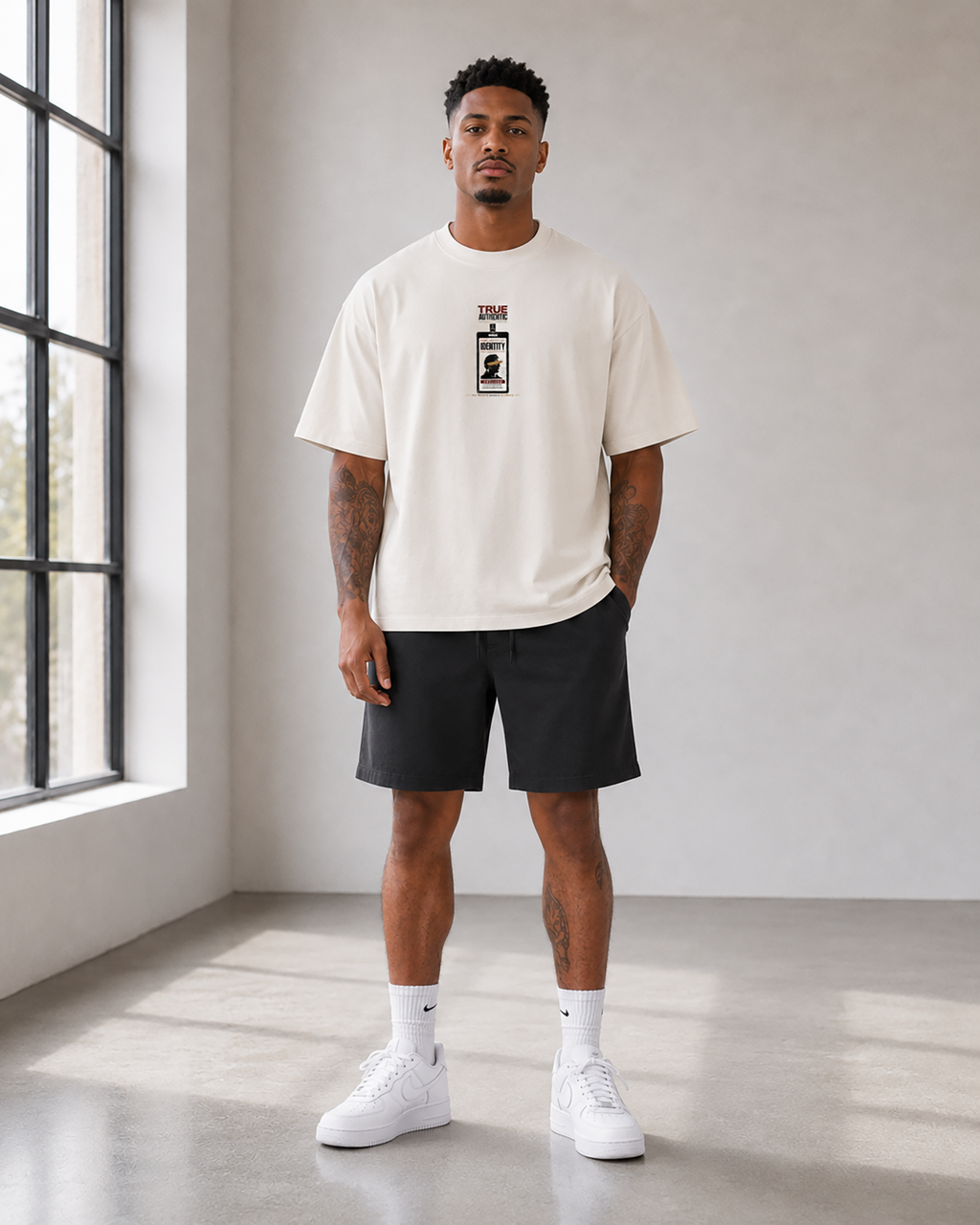 A man stands in a bright studio wearing an off-white t-shirt with a black graphic print on the chest, black shorts, white socks, and white sneakers.