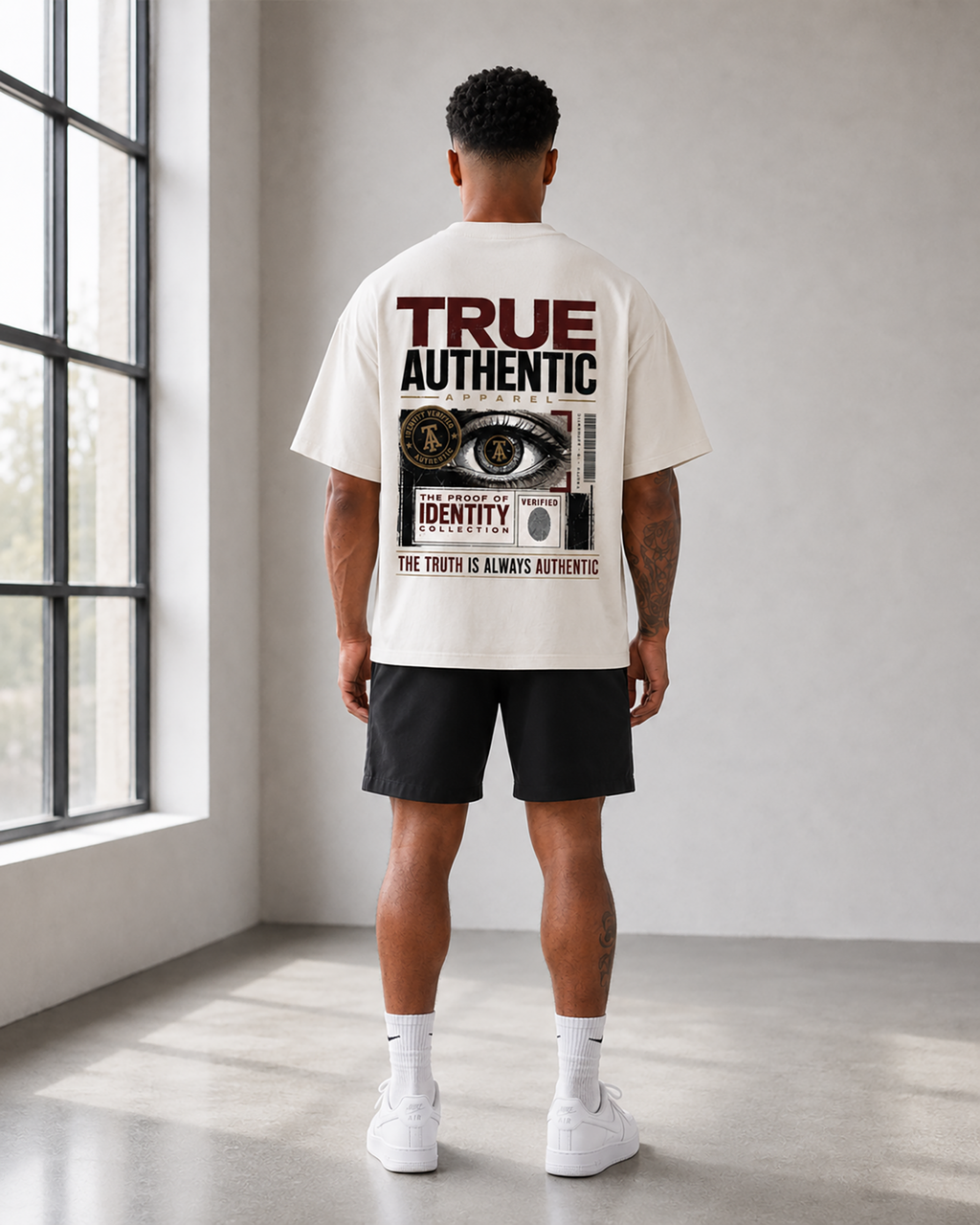 A man viewed from behind wearing an off-white graphic t-shirt with a large back print, black athletic shorts, white socks, and white sneakers in a bright, modern room with a large window.