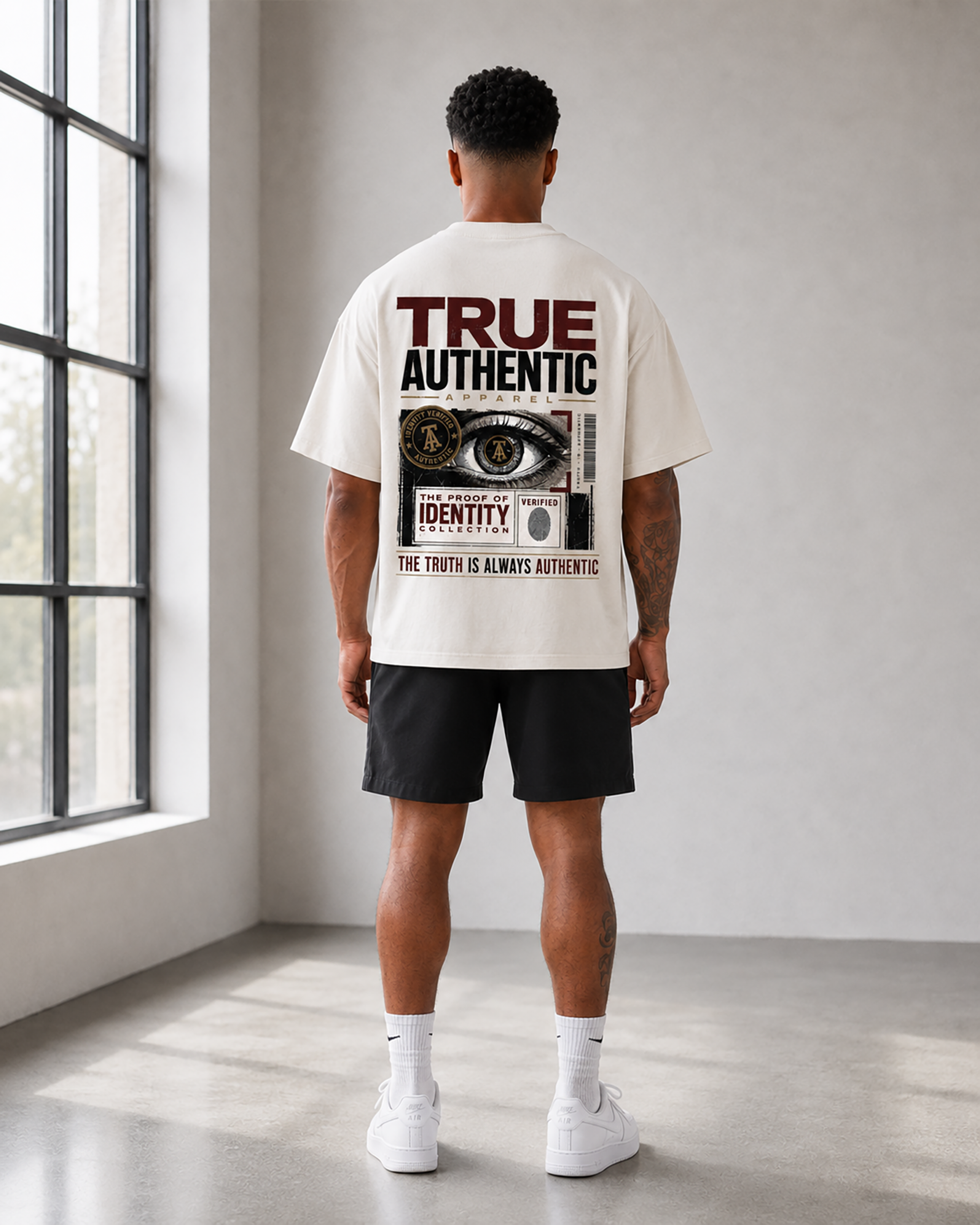A man viewed from behind wearing an off-white graphic t-shirt with a large back print, black athletic shorts, white socks, and white sneakers in a bright, modern room with a large window.