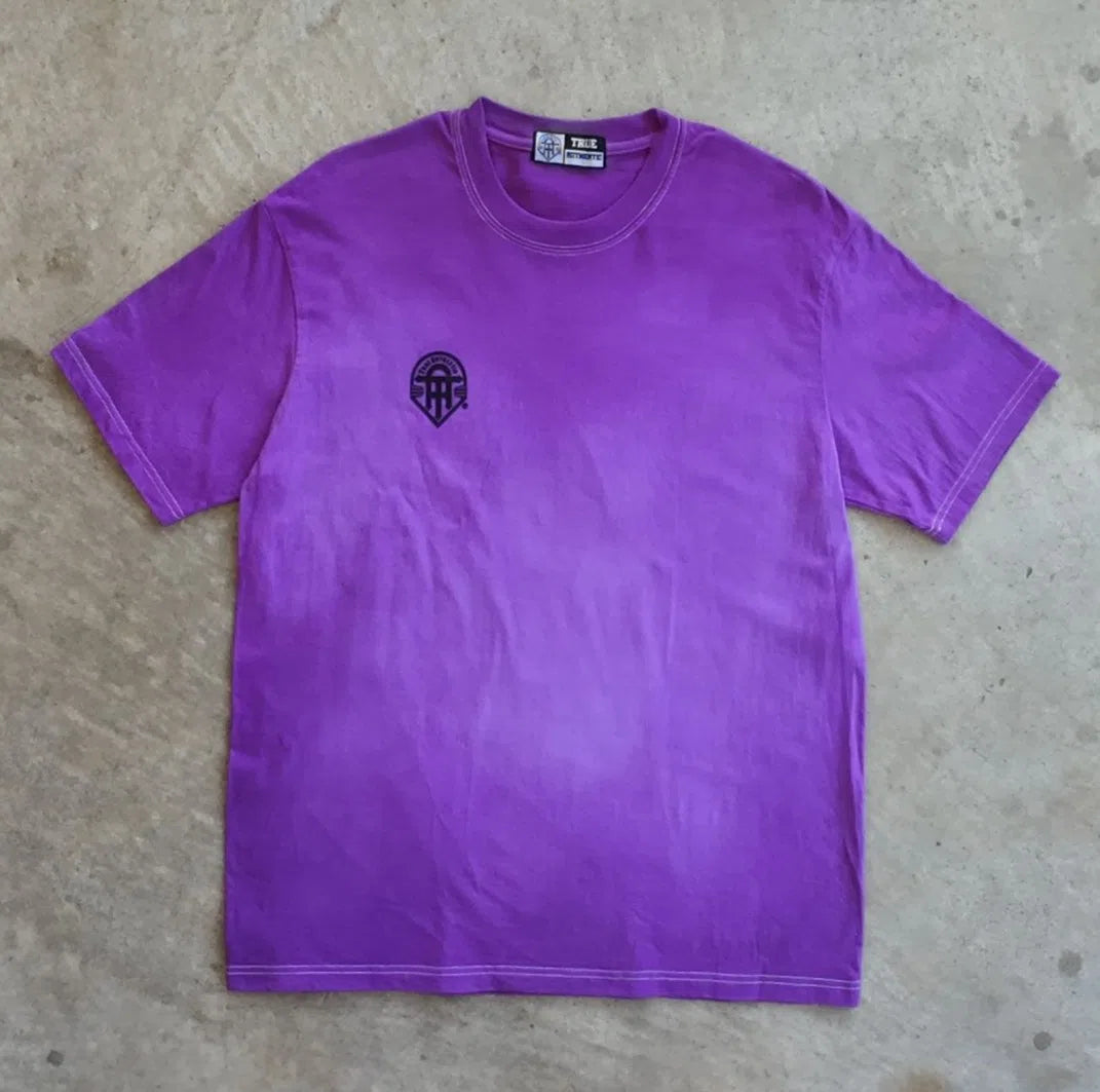 A purple t-shirt with a small logo on a concrete floor