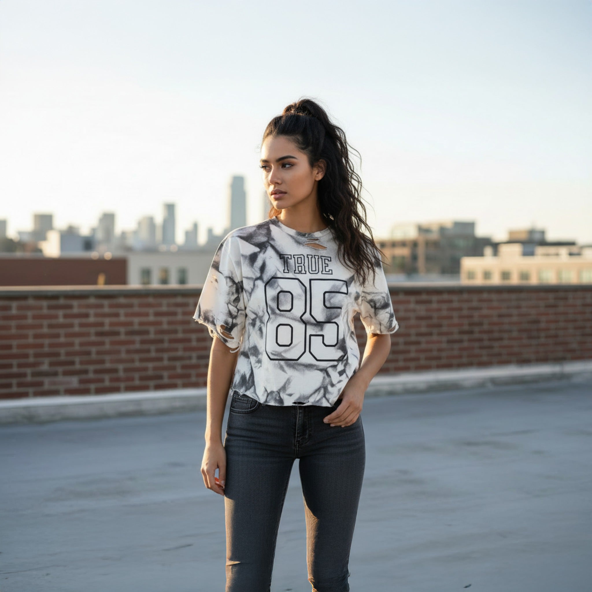 Woman wearing a tie-dye t-shirt with 'TRUE 85' on a rooftop with city skyline in the background