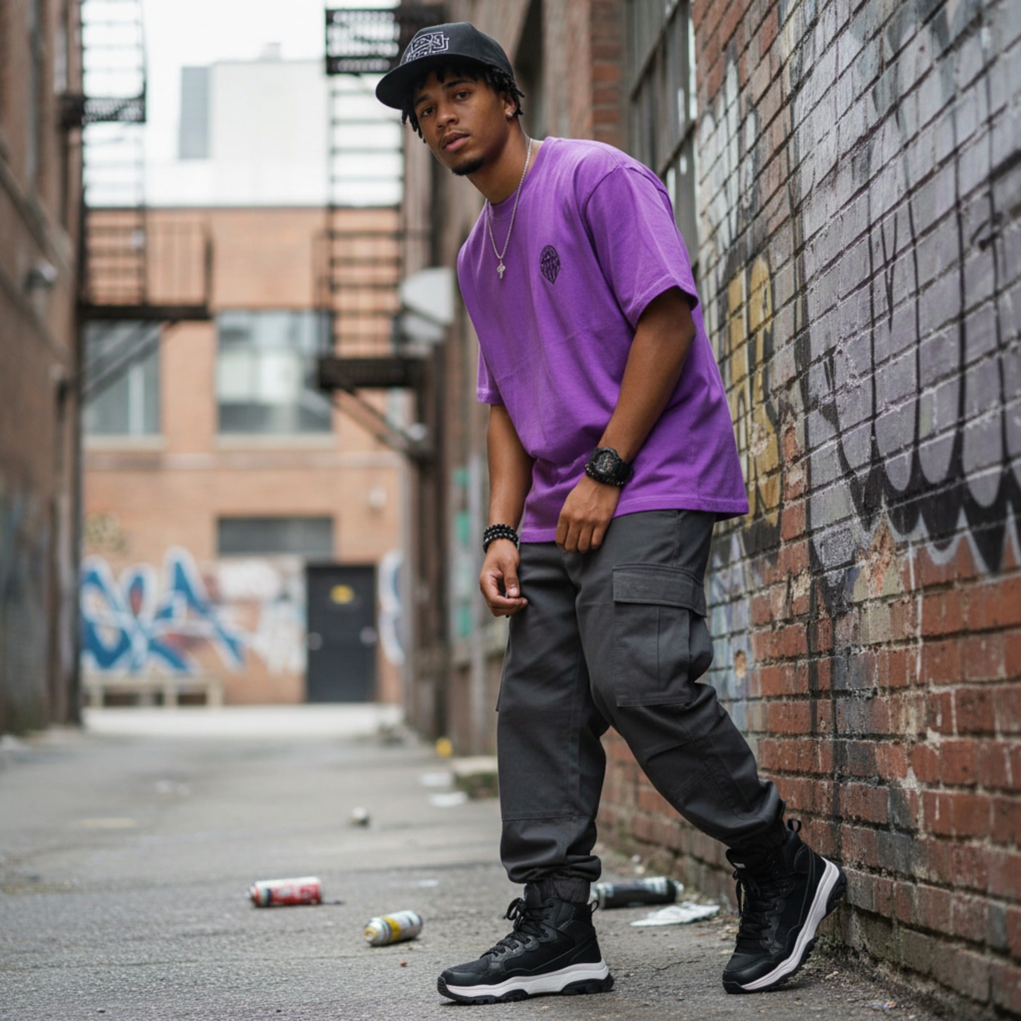 Person wearing a purple shirt and black pants leaning against a brick wall with graffiti.