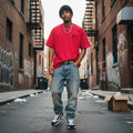 Man wearing a red t-shirt and blue jeans standing in an urban alleyway.